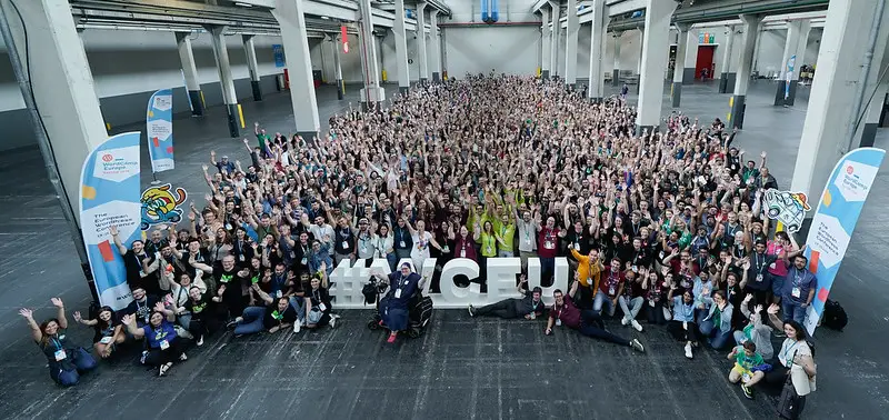 Foto di gruppo al WordCamp Europe 2024 di Torino. La foto è ripresa dall'alto, all'interno del padiglione 2 di Lingotto Fiere a Torino. In primo piano spicca la scritta grande in polistirolo bianco con l'hashtag #WCEU