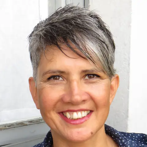 Foto in primo piano di Laura Sacco, capelli sale e pepe corti, con un ciuffo a sinistra un poco più lungo, labbra e occhi sorridenti